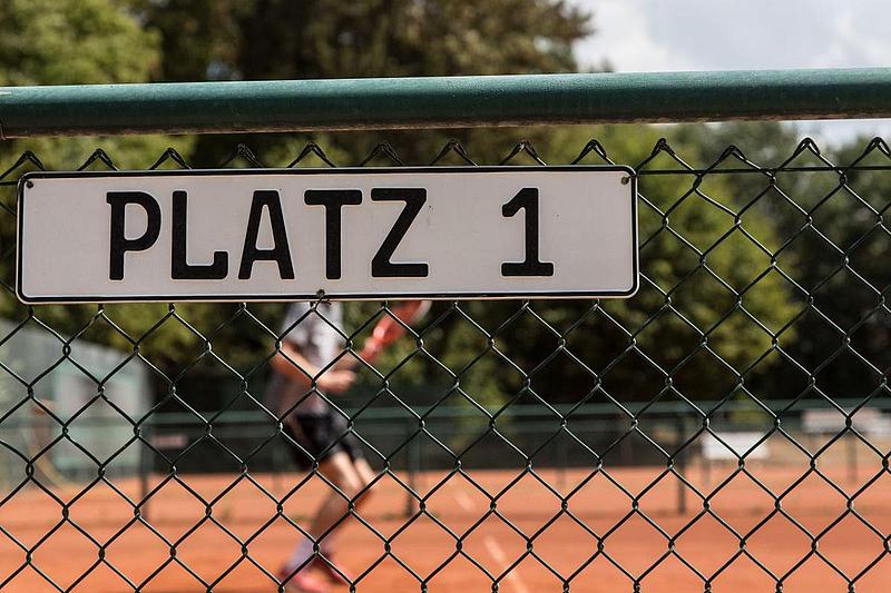Tennisplatz Nahaufnahme des Schildes "Platz 1", im Hintergrund ist ein Tennisspieler zu erkennen