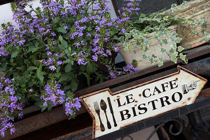 Nahaufnahme: Ein antikes Metallschild mit der Aufschrift "Le Cafe Bistro", an einem Metallgelände befestigt. Oberhalb des Geländes stehen Blumenkästen mit Blumen.