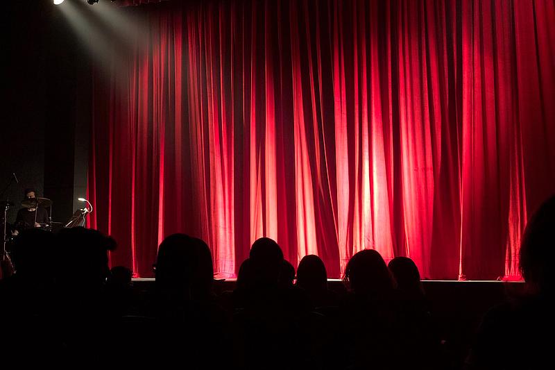 Publikum sitzt im Dunklen vor einem angestrahlten roten Vorhang